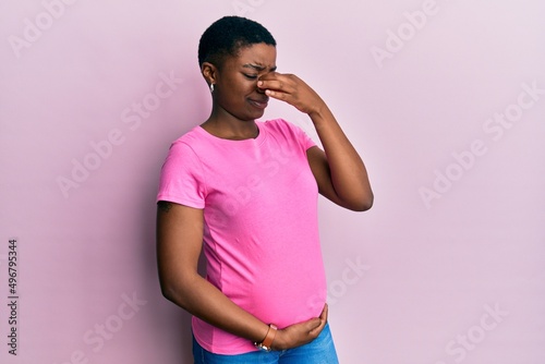 Young african american woman expecting a baby, touching pregnant belly smelling something stinky and disgusting, intolerable smell, holding breath with fingers on nose. bad smell