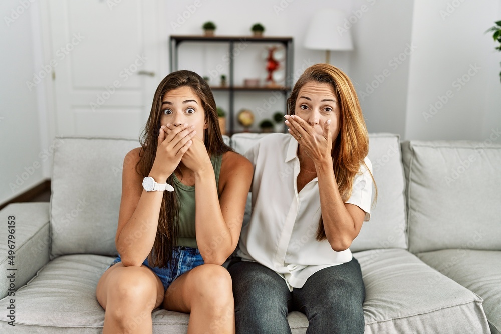 Mother and daughter together sitting on the sofa at home shocked covering mouth with hands for mistake. secret concept. Mother and daughter together sitting on the sofa at home shocked covering mouth with hands for mistake. secret concept.