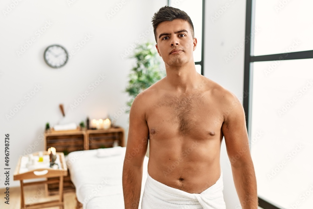 Obraz premium Young hispanic man standing shirtless at spa center relaxed with serious expression on face. simple and natural looking at the camera.