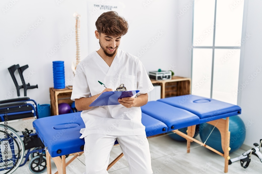 Young arab man wearing physiotherapist uniform writing on clipboard at clinic