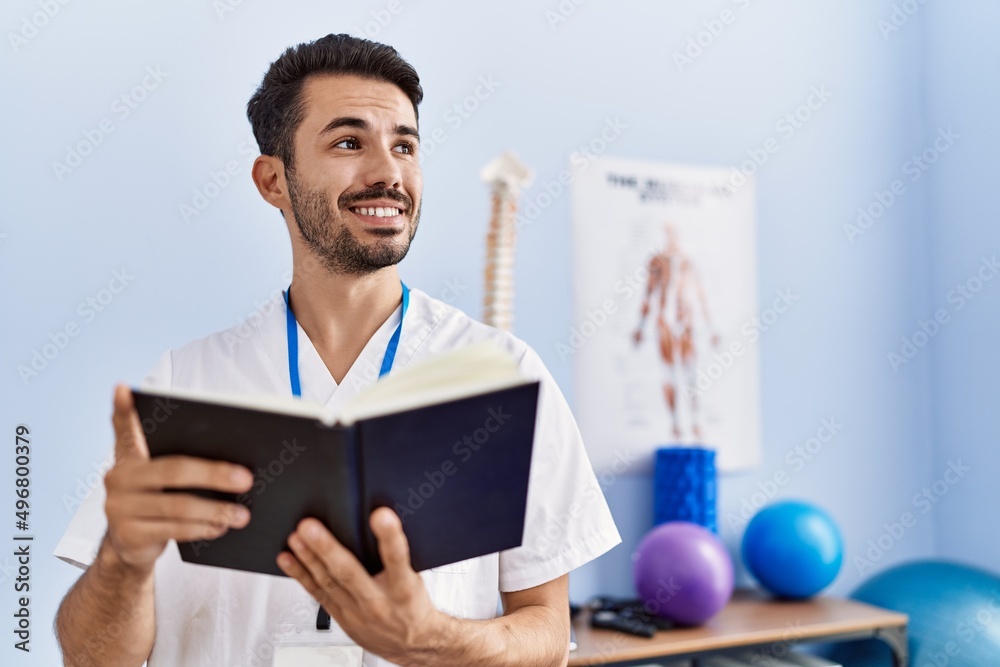 Young hispanic man wearing physiotherapist uniform reading book at rehab clinic Young hispanic man wearing physiotherapist uniform reading book at rehab clinic