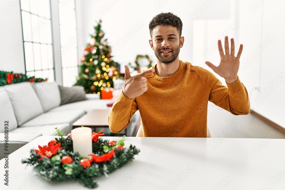 Arab young man sitting on the table by christmas tree showing and pointing up with fingers number seven while smiling confident and happy. Arab young man sitting on the table by christmas tree showing and pointing up with fingers number seven while smiling confident and happy.