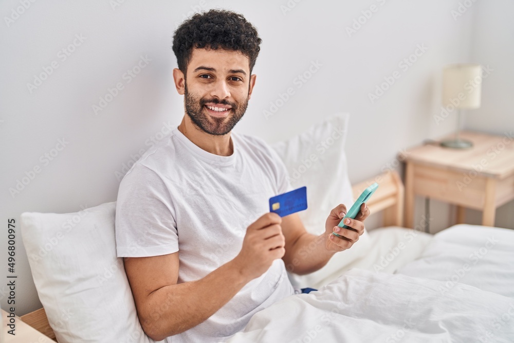 Young arab man using smartphone and credit card sitting on bed at bedroom Young arab man using smartphone and credit card sitting on bed at bedroom