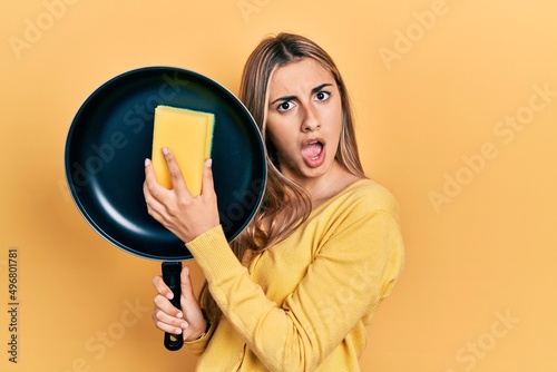 Beautiful hispanic woman cleaning cooking pan with scourer in shock face, looking skeptical and sarcastic, surprised with open mouth photo