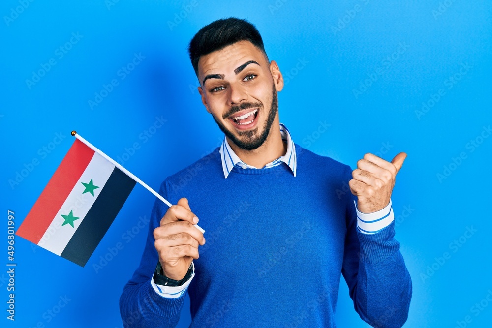 Young hispanic man with beard holding syria flag pointing thumb up to the side smiling happy with open mouth Young hispanic man with beard holding syria flag pointing thumb up to the side smiling happy with open mouth