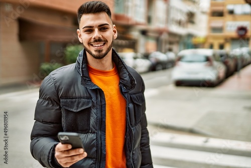 Handsome hispanic man with beard smiling happy and confident at the city wearing winter coat using smartphone