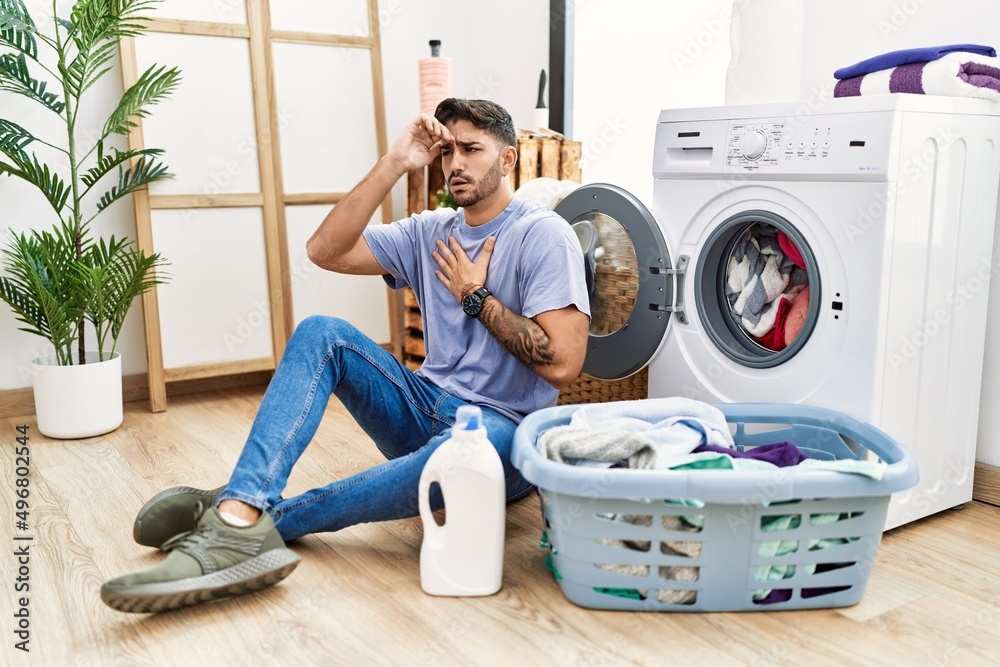 Young hispanic man putting dirty laundry into washing machine touching forehead for illness and fever, flu and cold, virus sick Young hispanic man putting dirty laundry into washing machine touching forehead for illness and fever, flu and cold, virus sick