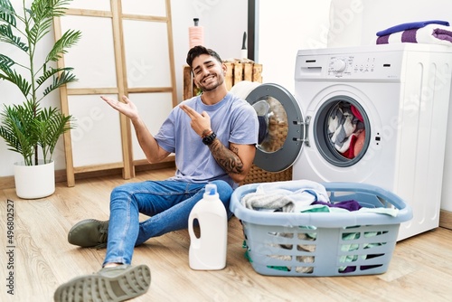 Young hispanic man putting dirty laundry into washing machine amazed and smiling to the camera while presenting with hand and pointing with finger.