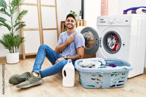 Young hispanic man putting dirty laundry into washing machine cheerful with a smile on face pointing with hand and finger up to the side with happy and natural expression