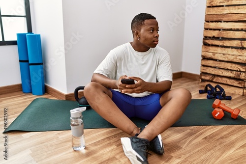 Young african man sitting on training mat at the gym using smartphone looking to side, relax profile pose with natural face and confident smile.