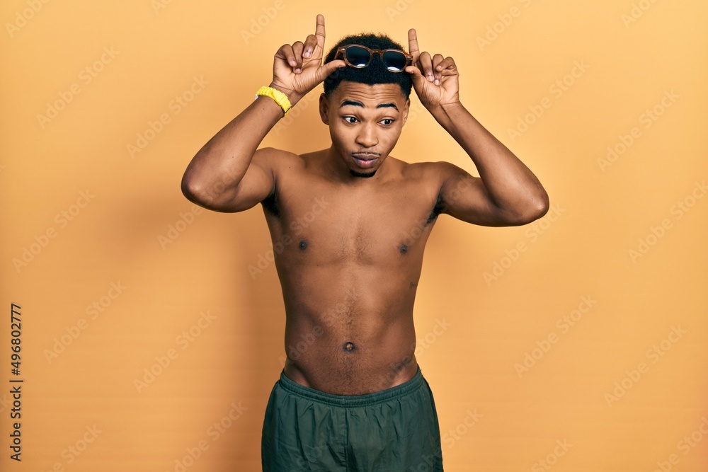 Young african american man wearing swimwear and swimmer glasses doing funny gesture with finger over head as bull horns