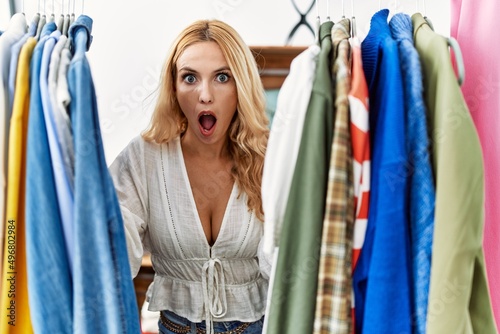 Beautiful blonde woman searching clothes on clothing rack afraid and shocked with surprise and amazed expression, fear and excited face.