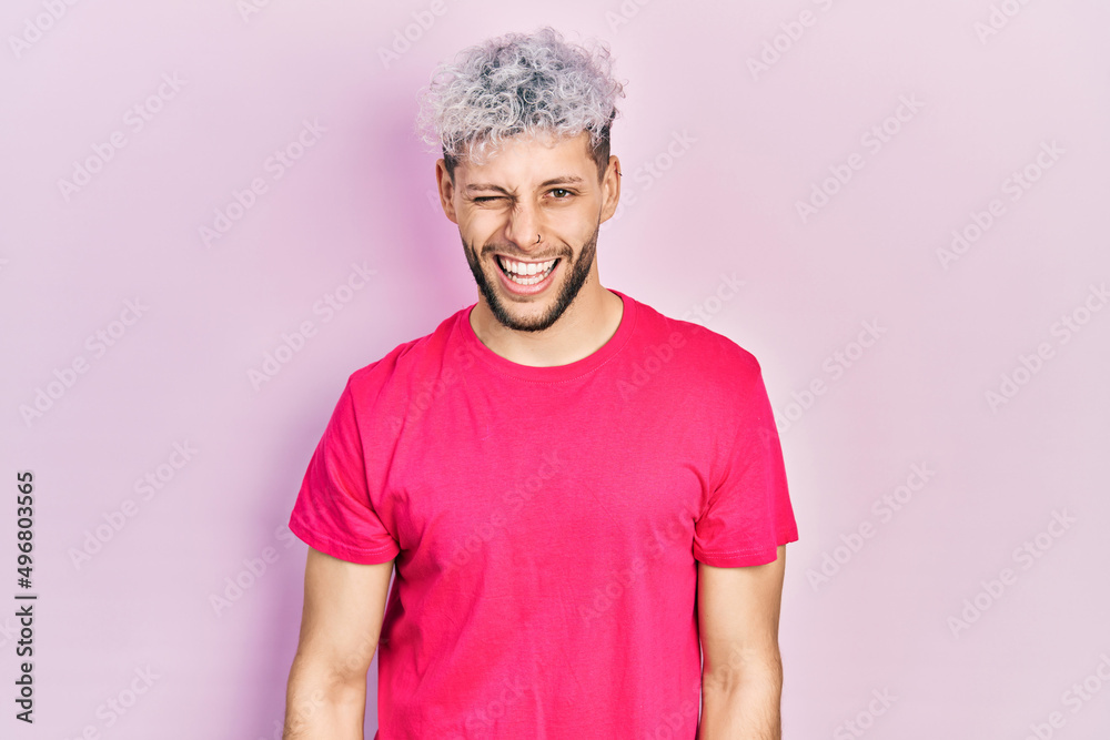 Young hispanic man with modern dyed hair wearing casual pink t shirt winking looking at the camera with sexy expression, cheerful and happy face. Young hispanic man with modern dyed hair wearing casual pink t shirt winking looking at the camera with sexy expression, cheerful and happy face.