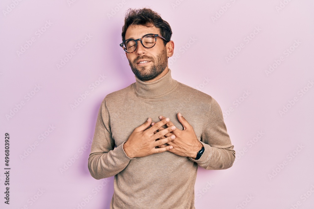 Handsome man with beard wearing turtleneck sweater and glasses smiling with hands on chest with closed eyes and grateful gesture on face. health concept. Handsome man with beard wearing turtleneck sweater and glasses smiling with hands on chest with closed eyes and grateful gesture on face. health concept.