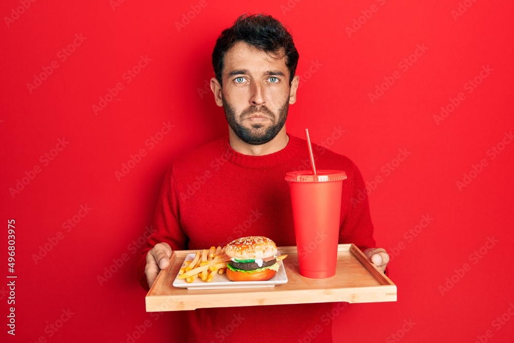 Handsome man with beard eating a tasty classic burger with fries and soda depressed and worry for distress, crying angry and afraid. sad expression. Handsome man with beard eating a tasty classic burger with fries and soda depressed and worry for distress, crying angry and afraid. sad expression.