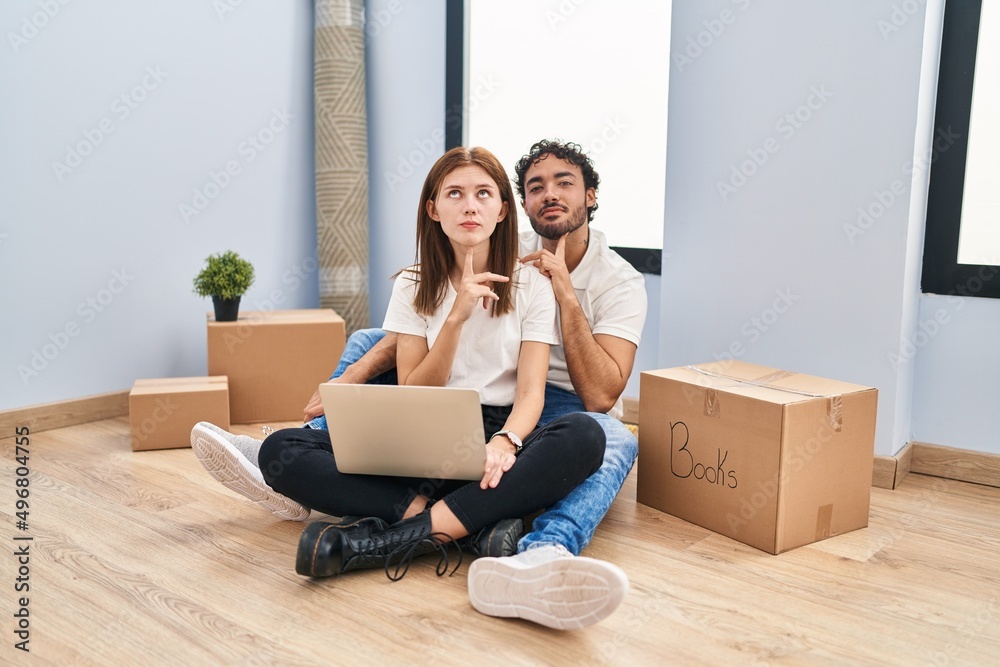 Young couple using laptop at new home thinking concentrated about doubt with finger on chin and looking up wondering Young couple using laptop at new home thinking concentrated about doubt with finger on chin and looking up wondering