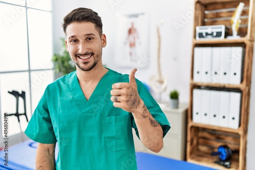 Young physiotherapist man working at pain recovery clinic doing happy thumbs up gesture with hand. approving expression looking at the camera showing success.