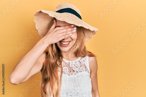 Young caucasian woman wearing summer hat smiling and laughing with hand on face covering eyes for surprise. blind concept.