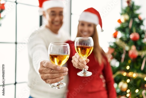 Middle age hispanic couple smiling happy drinking champagne standing by christmas tree at home.