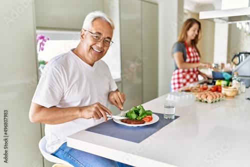 Middle age hispanic man smiling happy eating beef with salad while woman cook at the kitchen.