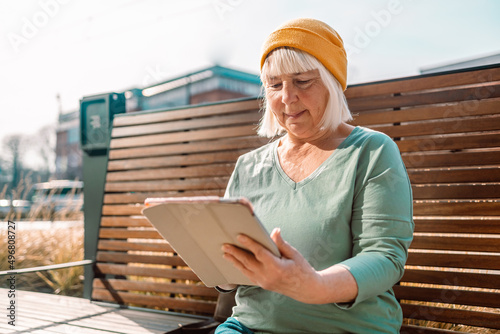 Fotografie 50s 60s Woman using digital tablet PC sitting on a modern wood bench in city wal
