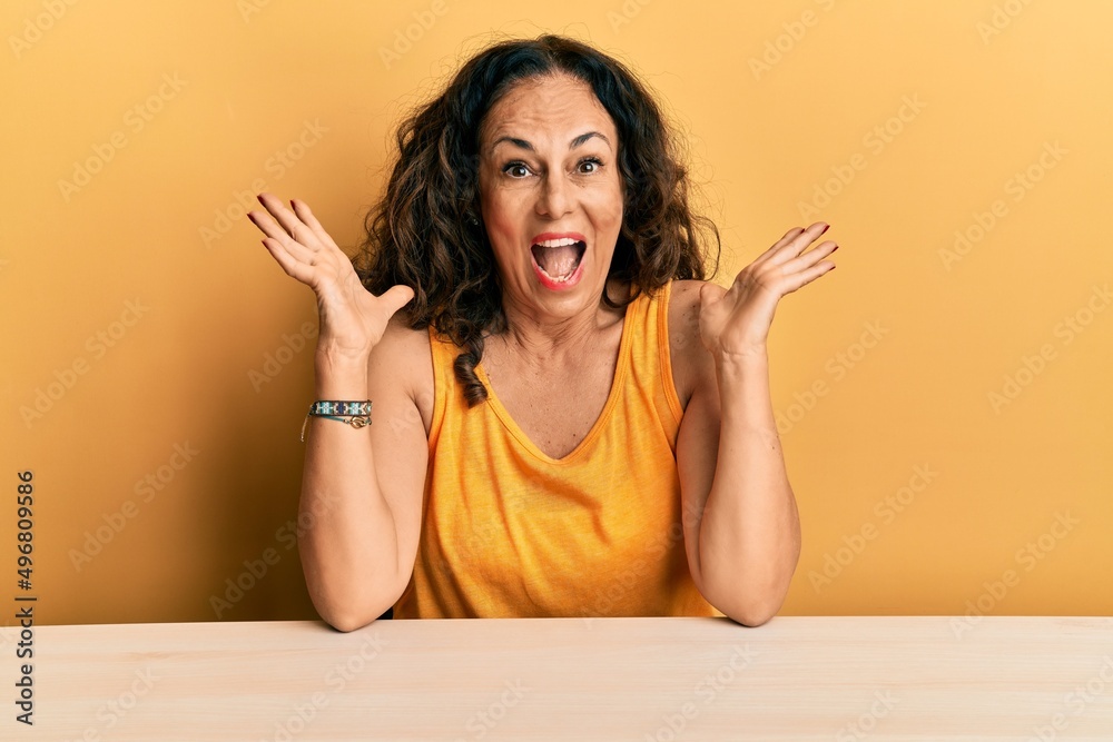 Beautiful middle age woman wearing casual clothes sitting on the table celebrating crazy and amazed for success with arms raised and open eyes screaming excited. winner concept Beautiful middle age woman wearing casual clothes sitting on the table celebrating crazy and amazed for success with arms raised and open eyes screaming excited. winner concept