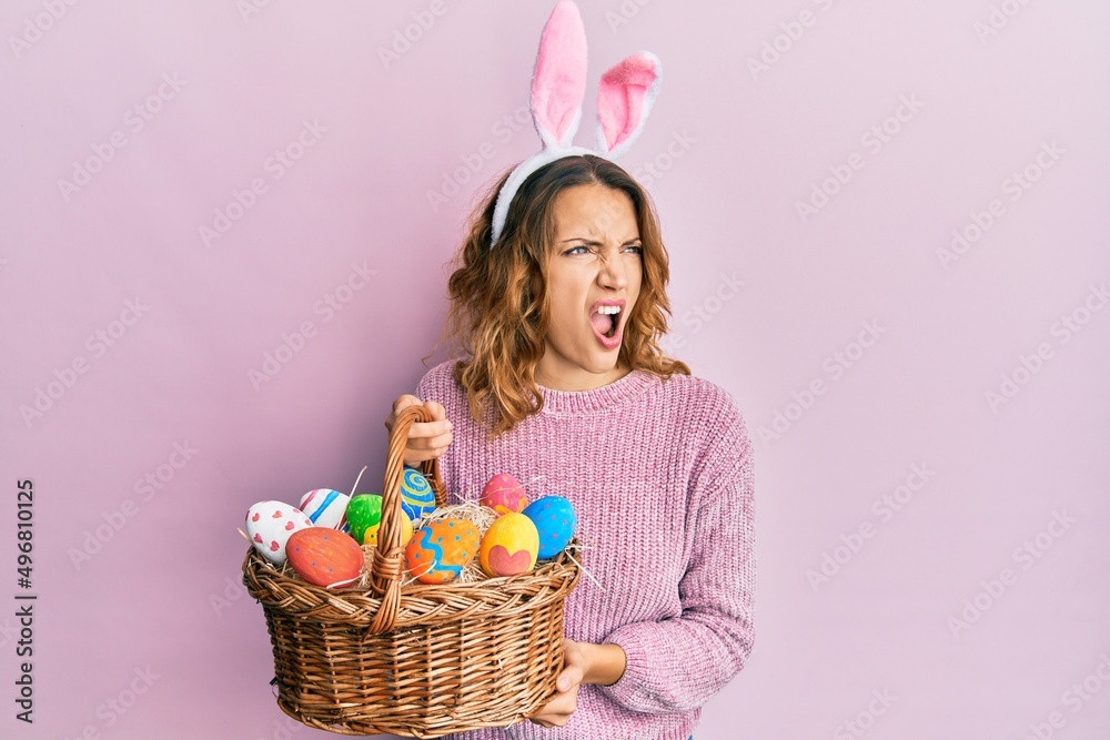 Young caucasian woman wearing cute easter bunny ears holding colored egg angry and mad screaming frustrated and furious, shouting with anger. rage and aggressive concept.