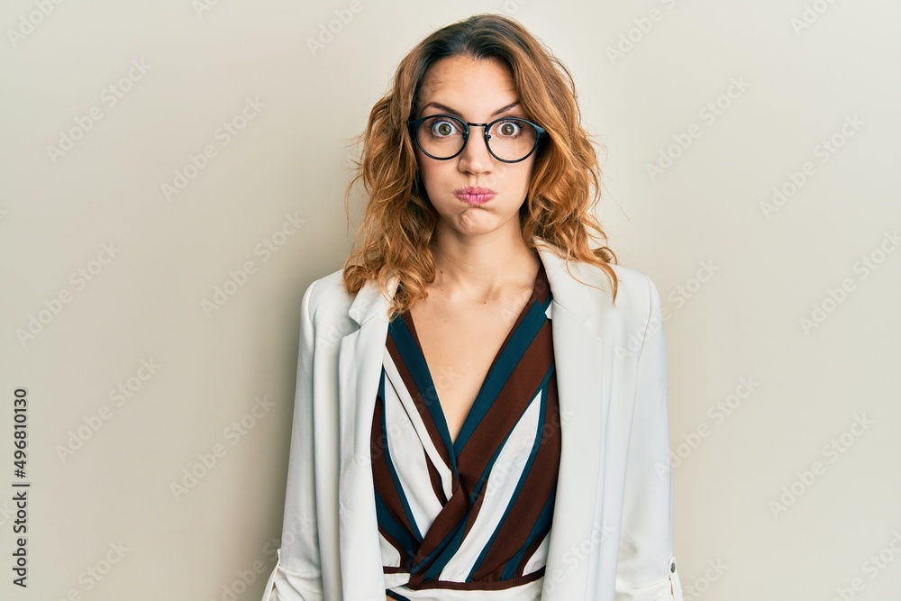 Young caucasian woman wearing business style and glasses puffing cheeks with funny face. mouth inflated with air, crazy expression.