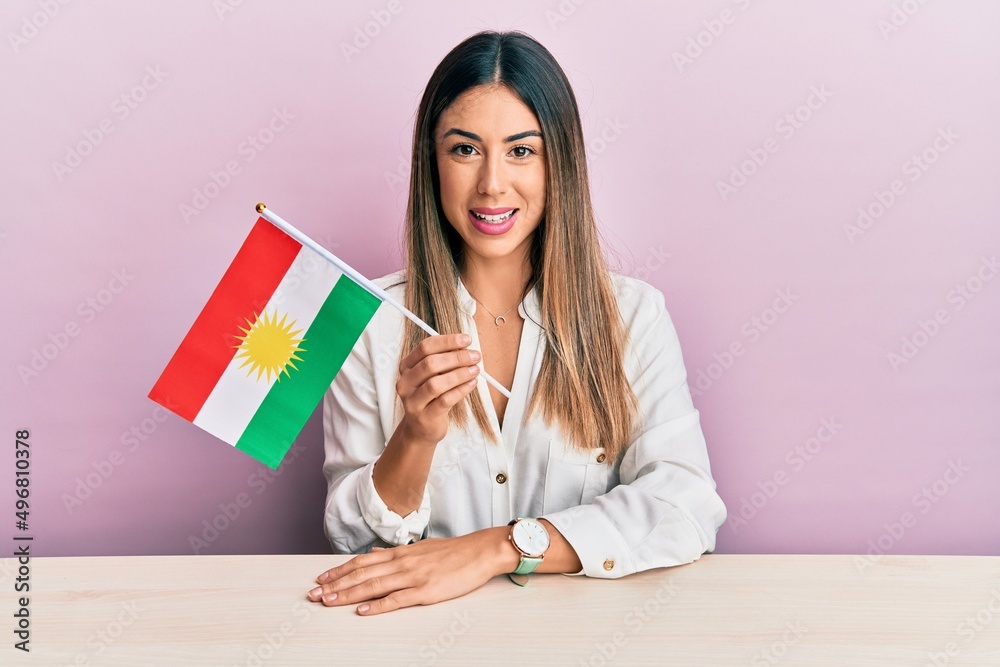 Young hispanic woman holding kurdistan flag sitting on the table looking positive and happy standing and smiling with a confident smile showing teeth