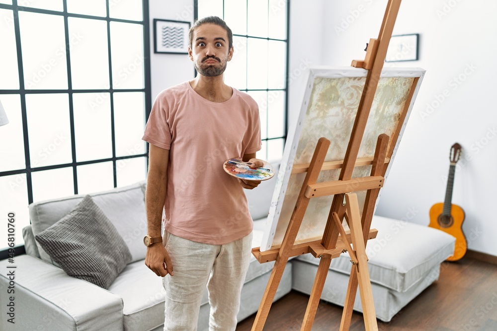 Young hispanic man with beard painting on canvas at home puffing cheeks with funny face. mouth inflated with air, crazy expression.