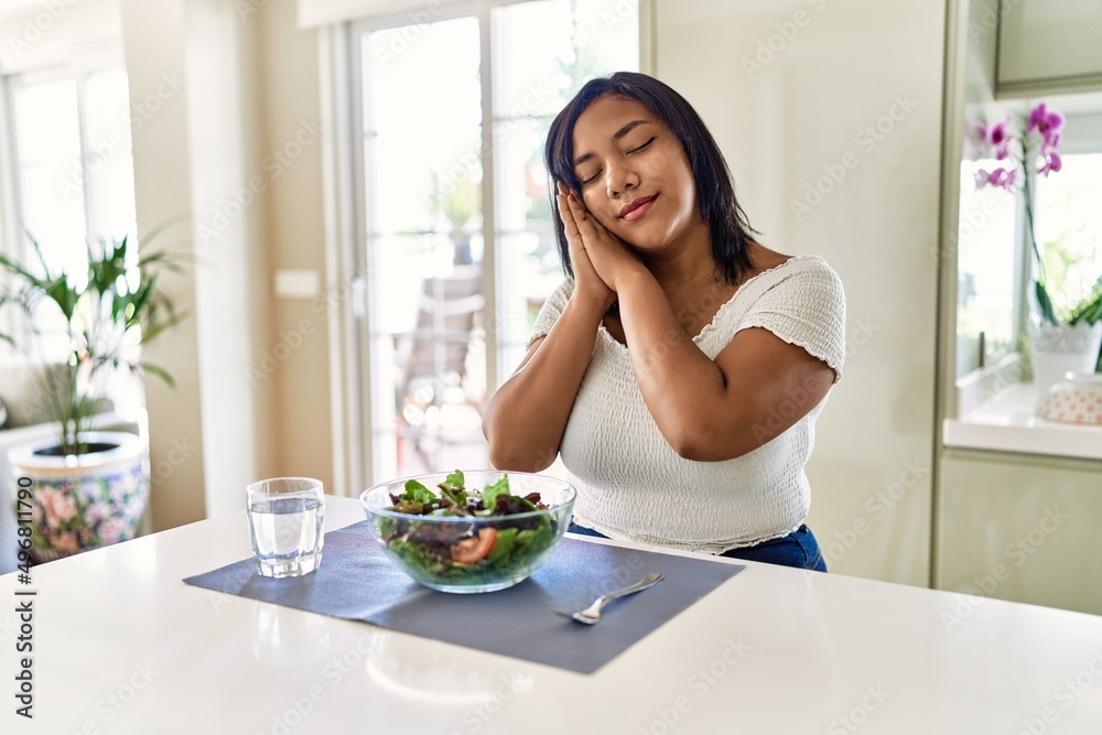 Young hispanic woman eating healthy salad at home sleeping tired dreaming and posing with hands together while smiling with closed eyes.