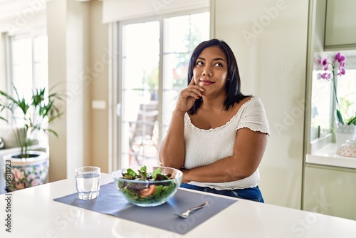 Young hispanic woman eating healthy salad at home with hand on chin thinking about question, pensive expression. smiling with thoughtful face. doubt concept.