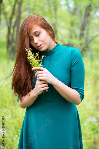 Redhead girl in forest on green trees background. Beautiful woman in elegant dress holds wildflowers in hands. Natural beauty concept.