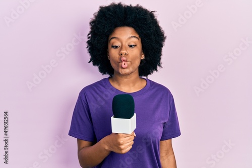 Young african american woman holding reporter microphone making fish face with mouth and squinting eyes, crazy and comical.