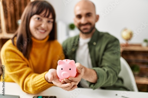 Young hispanic couple smiling happy holding piggy bank sitting on the table at home.