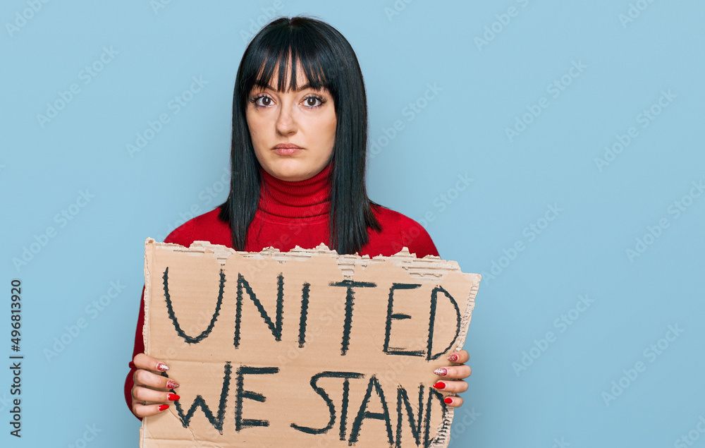 Young hispanic woman holding united we stand banner thinking attitude and sober expression looking self confident Young hispanic woman holding united we stand banner thinking attitude and sober expression looking self confident