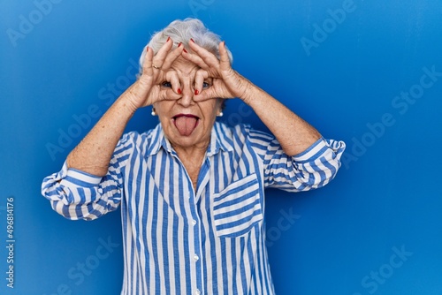 Senior woman with grey hair standing over blue background doing ok gesture like binoculars sticking tongue out, eyes looking through fingers. crazy expression.
