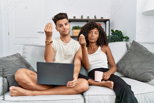 Young interracial couple using laptop at home sitting on the sofa doing italian gesture with hand and fingers confident expression