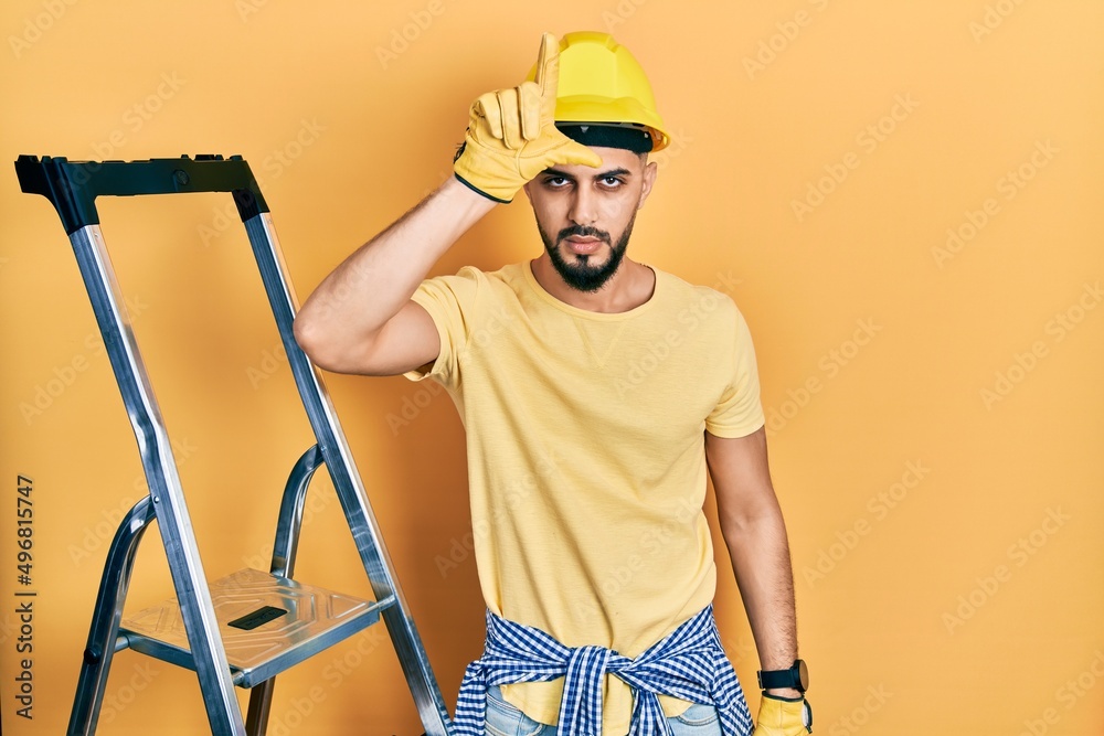 Handsome man with beard by construction stairs wearing hardhat making fun of people with fingers on forehead doing loser gesture mocking and insulting. Handsome man with beard by construction stairs wearing hardhat making fun of people with fingers on forehead doing loser gesture mocking and insulting.