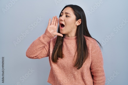 Young latin woman standing over blue background shouting and screaming loud to side with hand on mouth. communication concept.