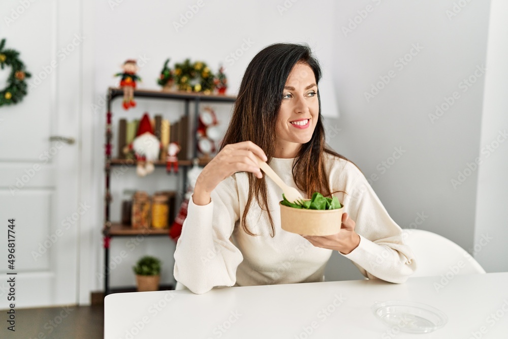 Middle age hispanic woman eating salad sitting on the table by christmas decor at home Middle age hispanic woman eating salad sitting on the table by christmas decor at home