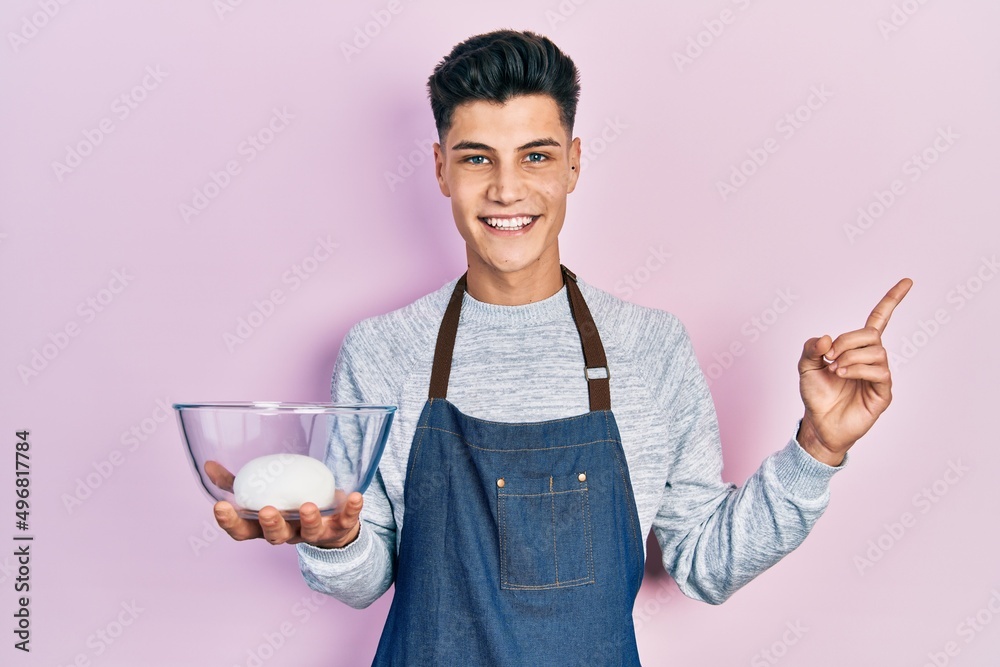 Young hispanic man wearing apron holding bowl of bread dough smiling happy pointing with hand and finger to the side Young hispanic man wearing apron holding bowl of bread dough smiling happy pointing with hand and finger to the side