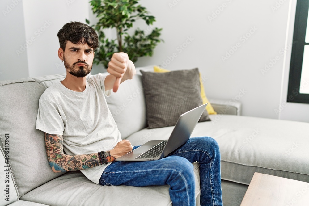 Hispanic man with beard sitting on the sofa looking unhappy and angry showing rejection and negative with thumbs down gesture. bad expression.