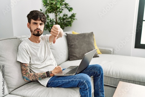 Hispanic man with beard sitting on the sofa looking unhappy and angry showing rejection and negative with thumbs down gesture. bad expression. © Krakenimages.com