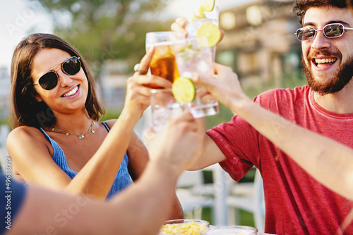 group of millenials toasting outdoors sitting on the terrace of a summer kiosk bar, young people friends having fun together drinking alcoholic drink cocktails on a sunny day and enjoying time