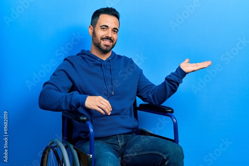 Handsome hispanic man with beard sitting on wheelchair smiling cheerful presenting and pointing with palm of hand looking at the camera.