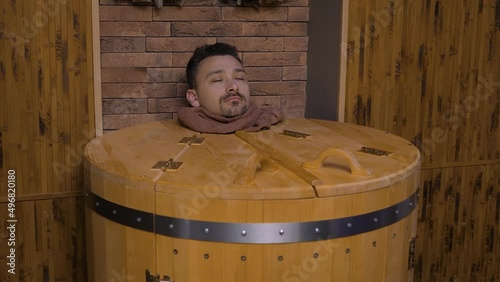 Male head peeks out of cedar hot herbal barrel at spa. Handsome middle-aged man receives sauna procedure with healing effect and enjoys the process of phyto barrel