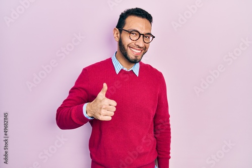 Hispanic man with beard wearing business shirt and glasses doing happy thumbs up gesture with hand. approving expression looking at the camera showing success.