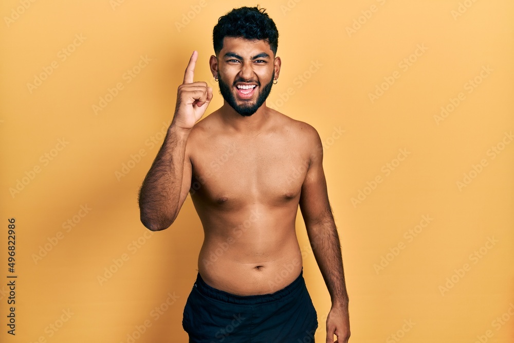 Arab man with beard wearing swimwear shirtless pointing finger up with successful idea. exited and happy. number one.