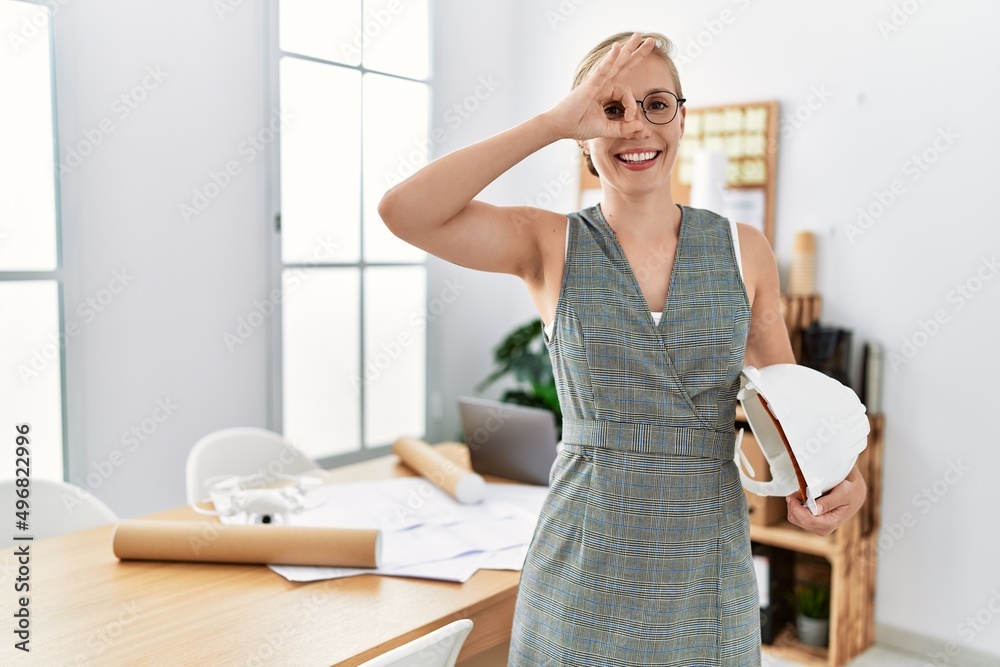 Young blonde woman holding architect hardhat smiling happy doing ok sign with hand on eye looking through fingers Young blonde woman holding architect hardhat smiling happy doing ok sign with hand on eye looking through fingers